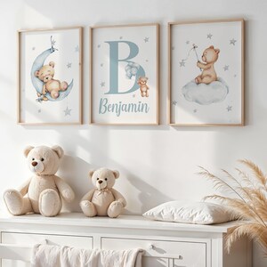 Könnte beinhalten: Drei gerahmte Kunstdrucke für das Kinderzimmer mit Aquarellillustrationen von Teddybären. Ein Druck zeigt einen Bären auf einem Mond, ein anderer den Buchstaben "B" und den Namen "Benjamin", und der dritte zeigt einen Bären auf einer Wolke. Zwei Teddybären-Plüschtiere stehen darunter.