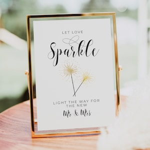 May include: A gold-framed sign with the text "Let love sparkle" in black cursive font. Two gold sparklers are depicted below the text. The text "Light the way for the new Mr. & Mrs." is printed below the sparklers.
