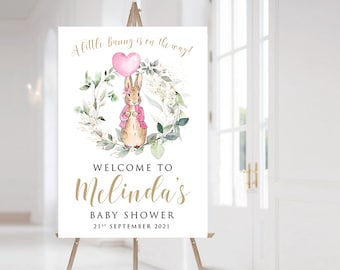 May include: A welcome sign for a baby shower with a watercolor illustration of a bunny holding a pink heart balloon. The sign says "A little bunny is on the way!" and "Welcome to Melinda's Baby Shower 21st September 2021".