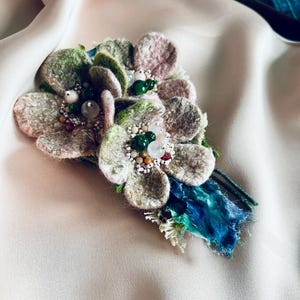 May include: A handmade floral brooch with felt petals in green, pink, and grey. The brooch is adorned with beads and green gemstones, and has a blue and green fabric accent.