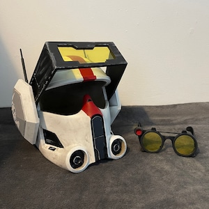 Op de afbeelding: Een witte en zwarte helm met een rode streep en een geel vizier. De helm heeft een zwart-wit zijpaneel met een klein wit schedelontwerp. Een paar zwarte brillen met gele glazen en een rode accent bevinden zich aan de rechterkant van de afbeelding.
