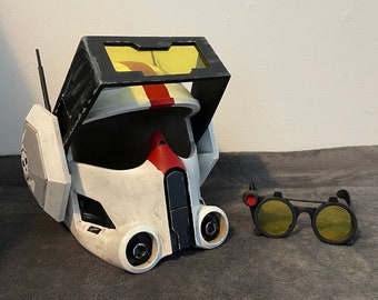 May include: A white and black helmet with a red stripe and yellow visor. The helmet has a black and white side panel with a small white skull design. A pair of black goggles with yellow lenses and a red accent are on the right side of the image.
