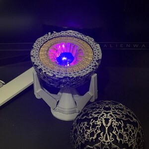 May include: A 3D printed model of a sphere with a detailed, intricate design. The sphere is open, revealing a glowing purple light inside. The sphere is on a stand with a base.