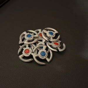 Puede incluir: Un montón de fichas de metal de color plateado con centros rojos y azules. Cada ficha tiene un diseño de tres puntas.