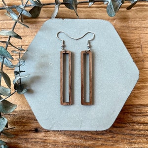 Puede incluir: Un par de pendientes largos, rectangulares de madera con un acabado de veta de madera natural. Los pendientes tienen un diseño simple y minimalista y están suspendidos de ganchos plateados.