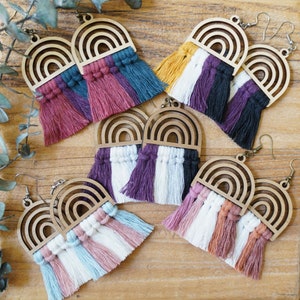May include: Wooden rainbow earrings with colorful macrame tassels. The earrings feature a laser-cut wooden rainbow design and are available in various color combinations, including pink, blue, and purple. The earrings have a hook closure.