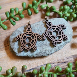 May include: A pair of wooden turtle earrings. The earrings are brown with a cut-out design, showcasing the turtle's shell and body. They are displayed on a gray stone, with green foliage in the background. The earrings have gold-colored hooks.