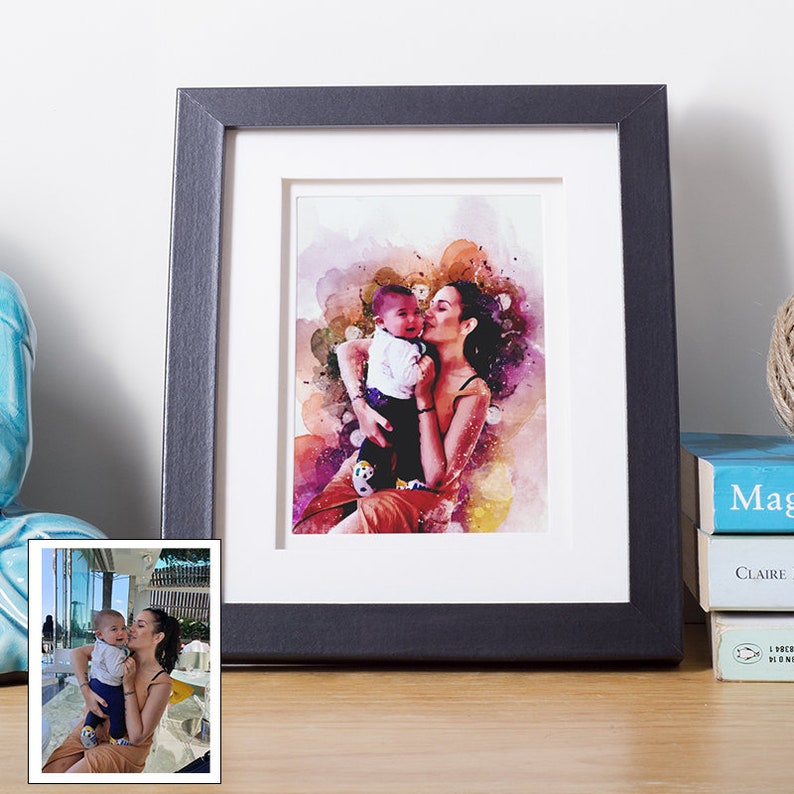 May include: A framed watercolor print of a mother and child, rendered in vibrant pink, purple, and orange hues. The black frame sits on a wooden surface, with a smaller photo of the same subjects in the lower left corner.
