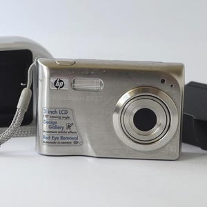 May include: Silver HP Photosmart R927 digital camera with a 3-inch LCD screen. The camera has a lens, flash, and a gray wrist strap. A silver case and a black charger are also visible.
