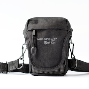 May include: A dark gray, rectangular camera bag with a shoulder strap and the text "ACROPOLIS series Uni-Set". The bag has a zippered main compartment and a small mesh pocket on the front. The strap is adjustable.