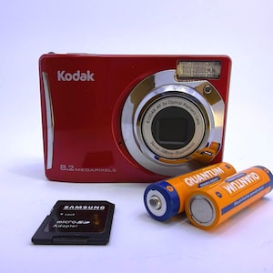 May include: A red Kodak digital camera with 8.2 megapixels and a 36mm-108mm lens, accompanied by two orange and blue Quantum batteries and a black Samsung microSD adapter. The camera features a silver lens housing and a built-in flash unit.