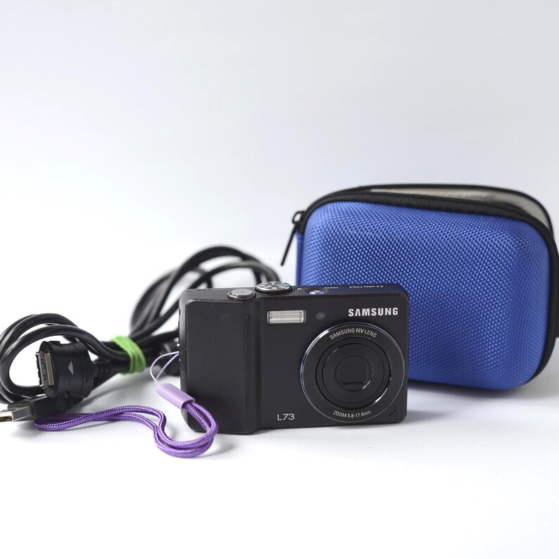 Puede incluir: C&aacute;mara digital Samsung L73 negra con una correa de mu&ntilde;eca morada, una funda de transporte azul y un cable de carga negro. La c&aacute;mara tiene un flash y una lente plateados. La funda es azul texturizada con cremallera negra.