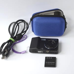 Puede incluir: Una c&aacute;mara digital Samsung NV7 negra con una funda azul, bater&iacute;a y cable de carga. La c&aacute;mara tiene el texto "L73" en la parte delantera. La bater&iacute;a tiene el texto "FOR DMW-BCB7/SD04E".