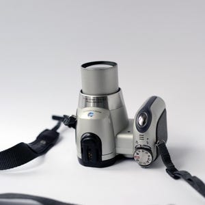 Pu&ograve; includere: Una fotocamera digitale argentata con una tracolla nera. La fotocamera ha un obiettivo zoom e una ghiera per regolare le impostazioni.