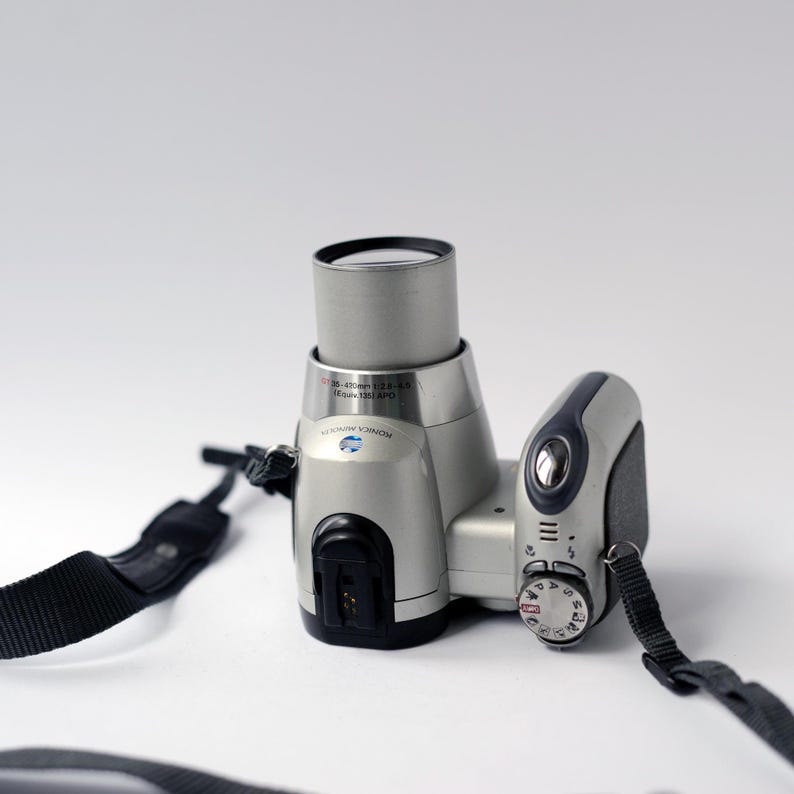 Pu&ograve; includere: Una fotocamera digitale argentata con una tracolla nera. La fotocamera ha un obiettivo zoom e una ghiera per regolare le impostazioni.