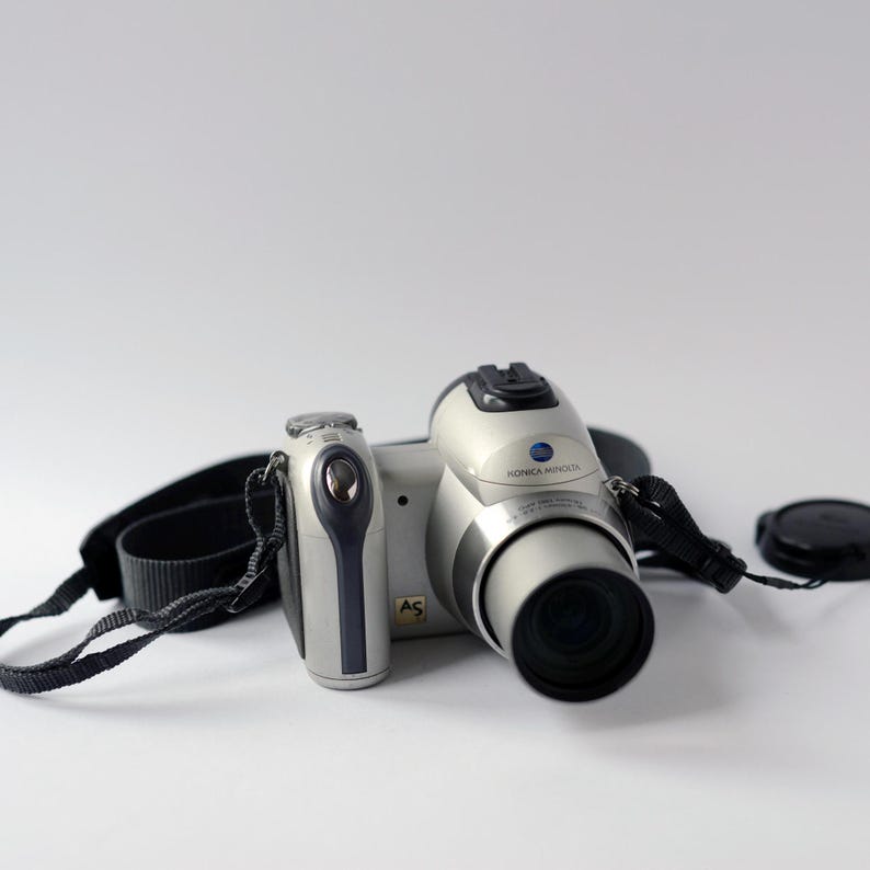 Pu&ograve; includere: Una fotocamera digitale Konica Minolta argentata con una tracolla e un tappo dell'obiettivo neri. La fotocamera ha un pulsante di zoom nero e un anello di zoom argentato.