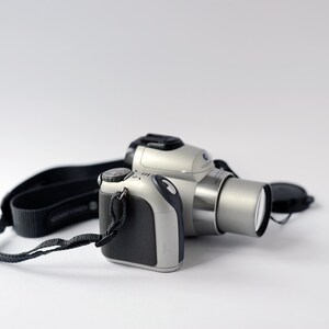 Pu&ograve; includere: Una fotocamera digitale argentata e nera con un copriobiettivo. La fotocamera ha una tracolla nera.