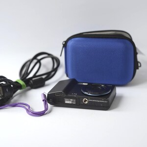 Puede incluir: Una c&aacute;mara digital negra con lente, una funda azul y un cable de carga negro con una correa de mu&ntilde;eca morada. La c&aacute;mara tiene el texto "L73" y "www.samsungcamera.com" impreso.