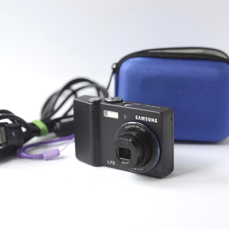 Puede incluir: C&aacute;mara digital Samsung L73 negra con un anillo de lente plateado y el logotipo de Samsung. Una funda azul para c&aacute;mara y un cable USB negro con una correa morada est&aacute;n en el fondo. La c&aacute;mara tiene un flash y una lente de 4,8-14,4 mm.