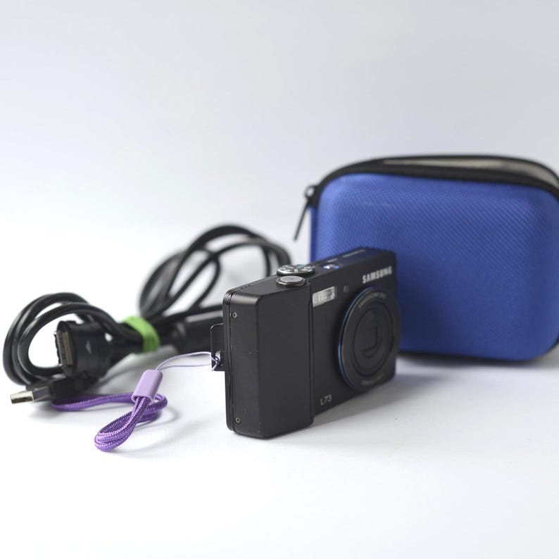 Puede incluir: Una c&aacute;mara digital Samsung L73 negra con una correa de mu&ntilde;eca morada, una funda de transporte azul y un cable USB negro. La c&aacute;mara est&aacute; sobre un fondo blanco.