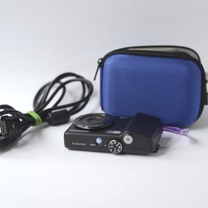 Puede incluir: Una c&aacute;mara digital Samsung negra con lente, bot&oacute;n de encendido y dial de control. Tambi&eacute;n hay una funda azul con cremallera y un cable de carga negro con una banda verde. La c&aacute;mara tiene impreso "7.2 MEGA PIXELS".