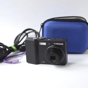 Puede incluir: C&aacute;mara digital Samsung L73 negra con una funda de transporte azul. La c&aacute;mara tiene una lente, flash y el logotipo de Samsung. Tambi&eacute;n se ve un cable de alimentaci&oacute;n con una correa de mu&ntilde;eca morada.