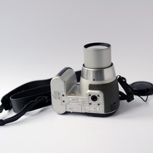 Pu&ograve; includere: Una fotocamera digitale argentata con una tracolla e un copriobiettivo neri. La fotocamera ha uno scomparto per batterie nero e un obiettivo argentato.