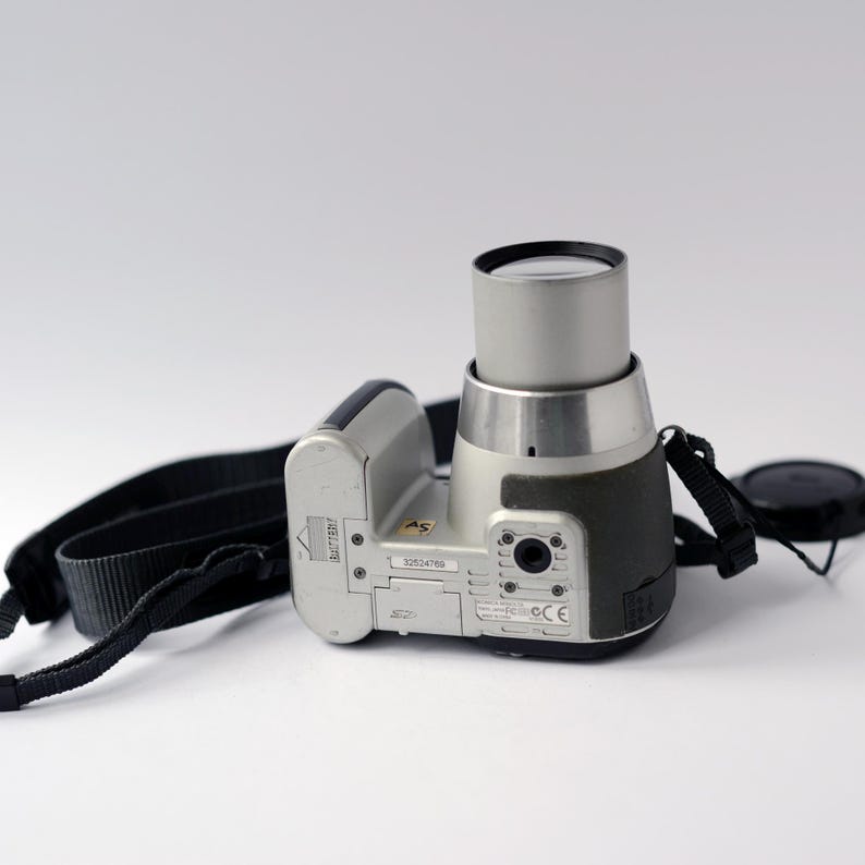 Pu&ograve; includere: Una fotocamera digitale argentata con una tracolla e un copriobiettivo neri. La fotocamera ha uno scomparto per batterie nero e un obiettivo argentato.
