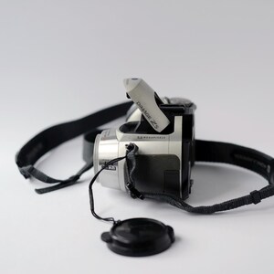 Pu&ograve; includere: Una fotocamera digitale argento e nera con una tracolla nera e un copriobiettivo. La fotocamera ha la scritta "DIMAGE Z5" e "5.0 MEGAPIXELS" stampata su di essa.