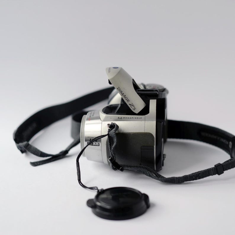 Pu&ograve; includere: Una fotocamera digitale argento e nera con una tracolla nera e un copriobiettivo. La fotocamera ha la scritta "DIMAGE Z5" e "5.0 MEGAPIXELS" stampata su di essa.
