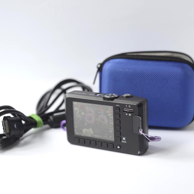 Puede incluir: Una c&aacute;mara digital negra con una pantalla que muestra una imagen floral, una funda de transporte azul y un cable de carga negro con una atadura verde. La c&aacute;mara tiene botones y una correa de mu&ntilde;eca morada.