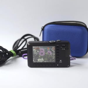 Puede incluir: Una c&aacute;mara digital negra con una pantalla que muestra una foto de flores moradas. Una funda azul con cremallera y un cable de alimentaci&oacute;n negro est&aacute;n detr&aacute;s de la c&aacute;mara. La c&aacute;mara tiene botones y una peque&ntilde;a pantalla. La imagen est&aacute; sobre un fondo blanco.