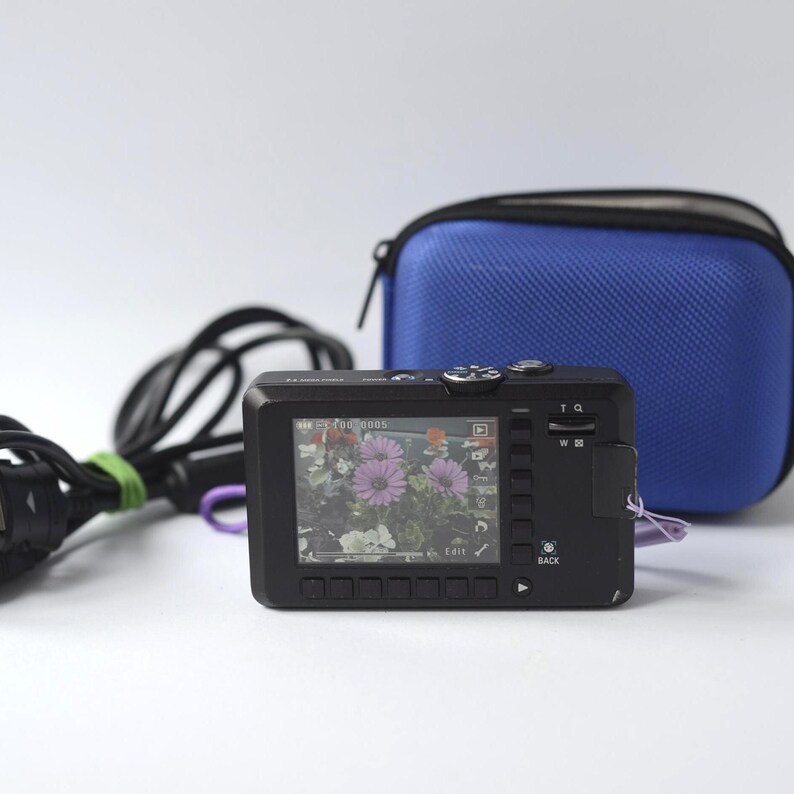 Puede incluir: Una c&aacute;mara digital negra con una pantalla que muestra una foto de flores moradas. Una funda azul con cremallera y un cable de alimentaci&oacute;n negro est&aacute;n detr&aacute;s de la c&aacute;mara. La c&aacute;mara tiene botones y una peque&ntilde;a pantalla. La imagen est&aacute; sobre un fondo blanco.