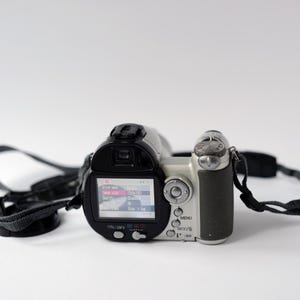 Pu&ograve; includere: Una videocamera digitale argento e nera con un cinturino nero. La videocamera ha un piccolo schermo LCD che mostra "Modalit&agrave; file", "Dimensione immagine", "Qualit&agrave;", "Zoom" e "Su/Gi&ugrave;/Cappuccio".