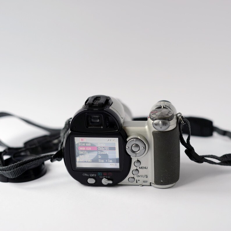 Pu&ograve; includere: Una videocamera digitale argento e nera con un cinturino nero. La videocamera ha un piccolo schermo LCD che mostra "Modalit&agrave; file", "Dimensione immagine", "Qualit&agrave;", "Zoom" e "Su/Gi&ugrave;/Cappuccio".
