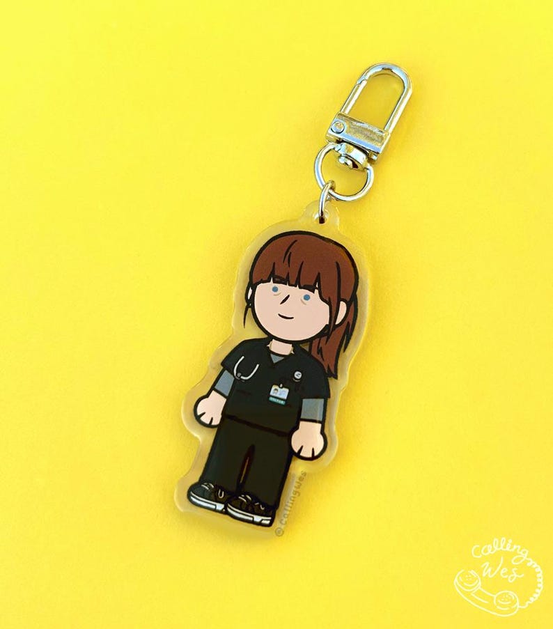 May include: A clear acrylic keychain featuring a cartoon illustration of a healthcare worker in black scrubs, a stethoscope, and brown hair. The keychain has a silver clasp and is set against a bright yellow background. The text "Calling Wes" is visible.