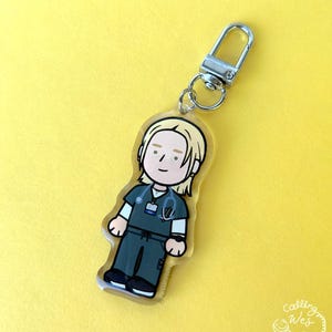 May include: A clear acrylic keychain featuring a cartoon illustration of a healthcare worker in teal scrubs, a stethoscope, and a name tag. The keychain has a silver clasp and a small ring. The background is a solid yellow.