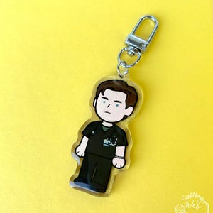 May include: A clear acrylic keychain featuring a cartoon illustration of a person in dark green scrubs. The keychain has a silver-colored clasp and a small ring. The background is a solid yellow color. The keychain has the text "Calling Wes" in the bottom right corner.