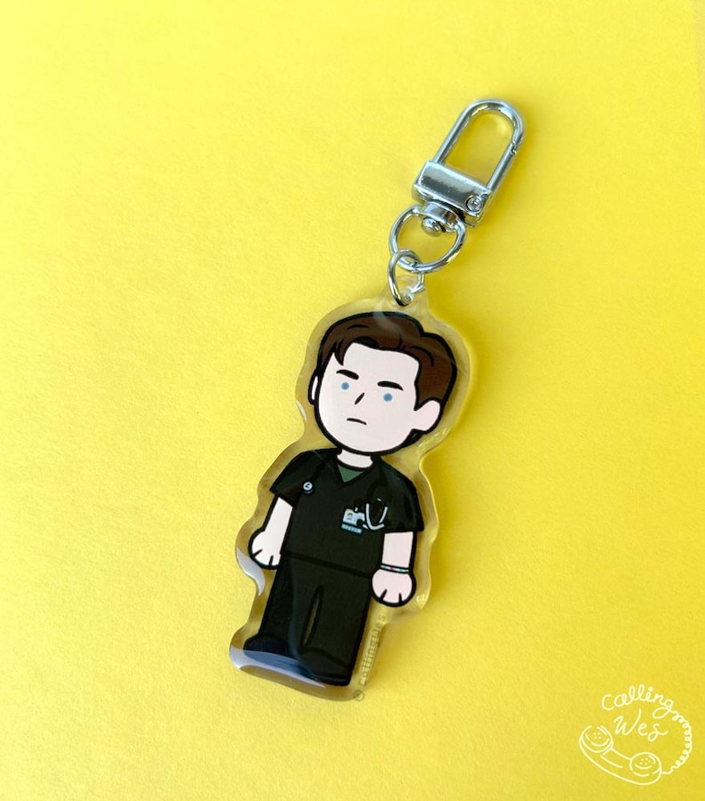 May include: A clear acrylic keychain featuring a cartoon illustration of a person in dark green scrubs. The keychain has a silver-colored clasp and a small ring. The background is a solid yellow color. The keychain has the text "Calling Wes" in the bottom right corner.