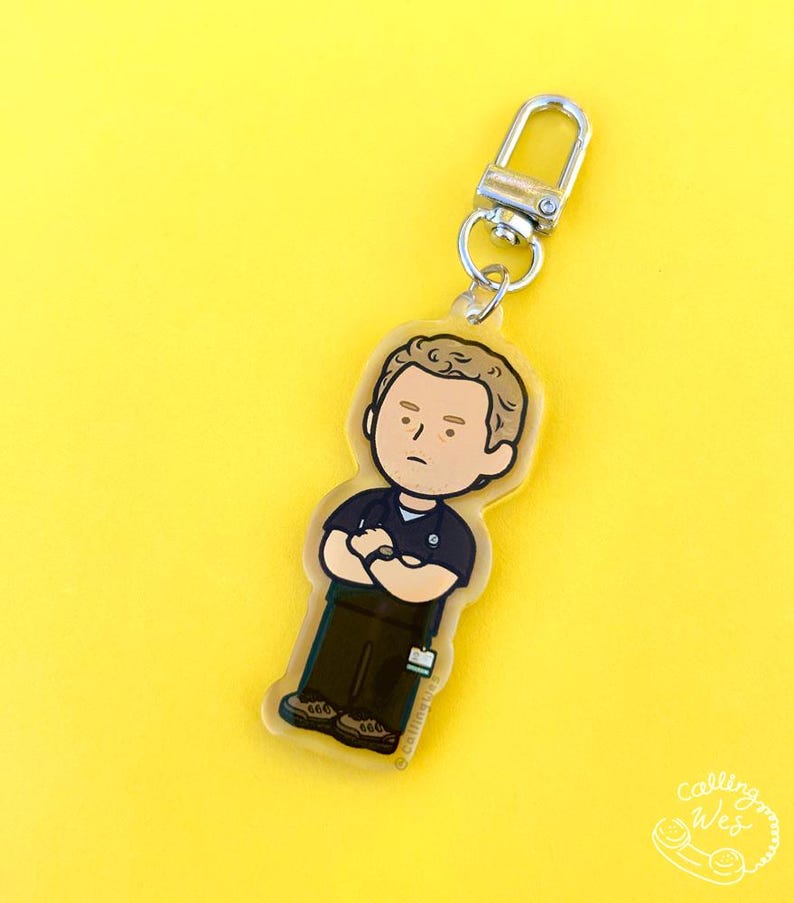 May include: A clear acrylic keychain featuring a cartoon illustration of a person in a dark blue shirt and brown pants with arms crossed. The keychain has a silver clasp and a small tag. The background is yellow.