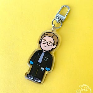 May include: A clear acrylic keychain featuring a cartoon illustration of a medical professional. The figure wears glasses, a black jacket, black pants, and blue gloves. The keychain has a silver clasp and hangs against a yellow background.