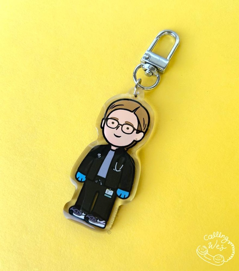 May include: A clear acrylic keychain featuring a cartoon illustration of a medical professional. The figure wears glasses, a black jacket, black pants, and blue gloves. The keychain has a silver clasp and hangs against a yellow background.