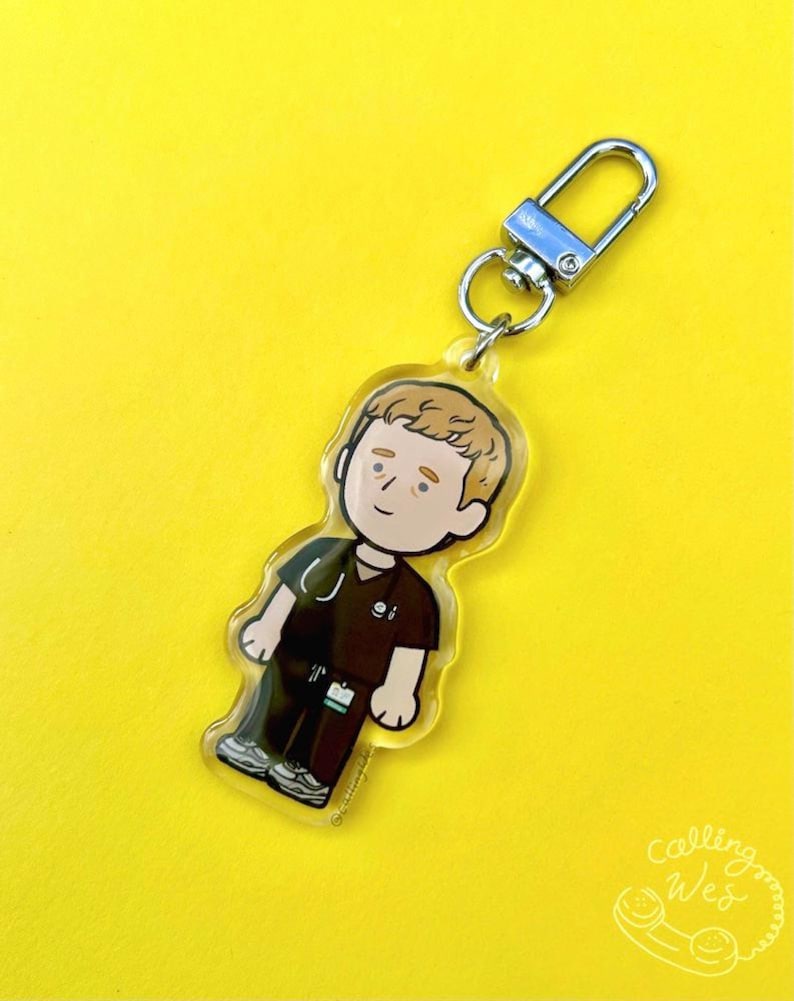 May include: A clear acrylic keychain featuring a cartoon illustration of a person in a dark brown medical uniform with a stethoscope. The keychain has a silver clasp and a small ring. The background is a bright yellow.