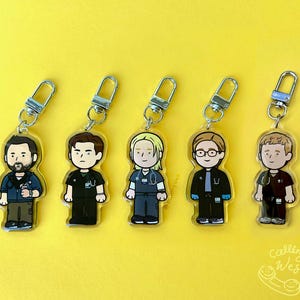 May include: Five acrylic keychains featuring cartoon illustrations of medical professionals. Each keychain has a silver clasp and depicts a different character in scrubs and a lab coat. The background is a solid yellow.