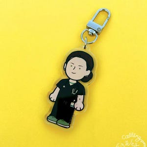 May include: A clear acrylic keychain featuring a cartoon illustration of a person in medical scrubs. The figure has dark hair and is wearing a black uniform with a green collar and a stethoscope. The keychain has a silver clasp and a yellow background.