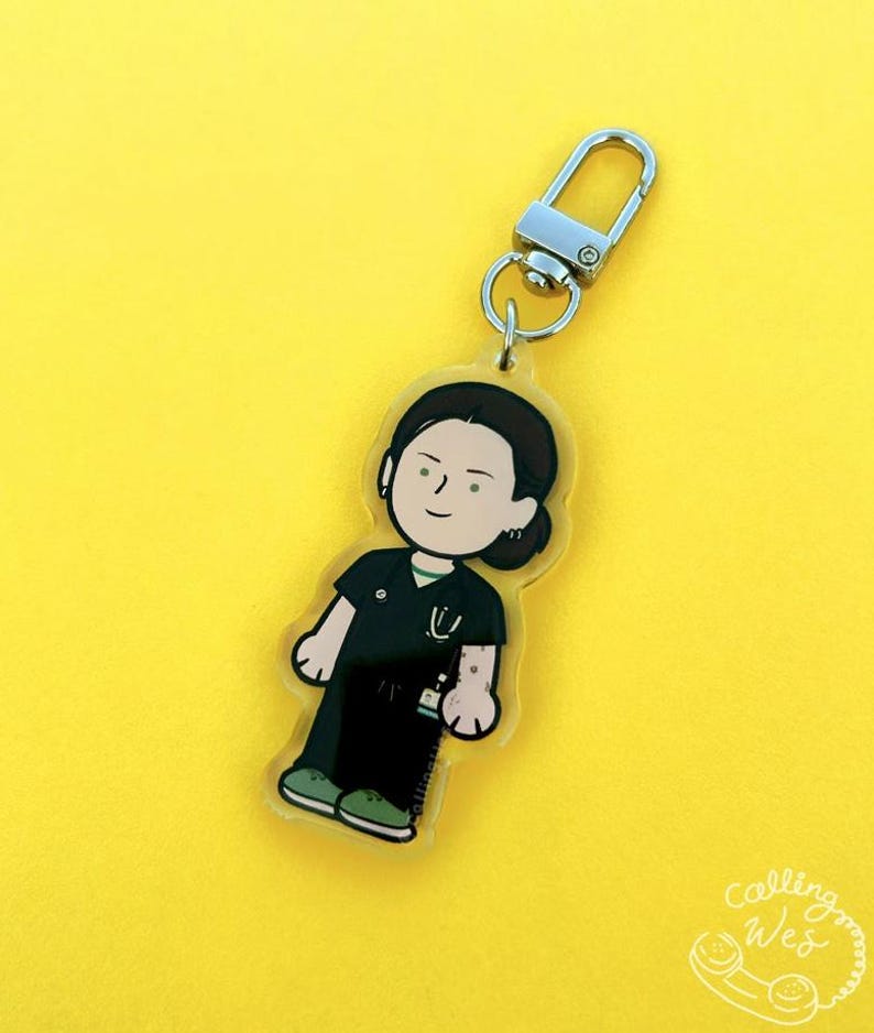 May include: A clear acrylic keychain featuring a cartoon illustration of a person in medical scrubs. The figure has dark hair and is wearing a black uniform with a green collar and a stethoscope. The keychain has a silver clasp and a yellow background.