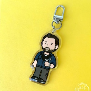 May include: A clear acrylic keychain featuring a cartoon illustration of a man with a beard, wearing a blue jacket, black shirt, and gray pants. The keychain has a silver clasp and a small coffee cup. The text "@callingwes" is visible.