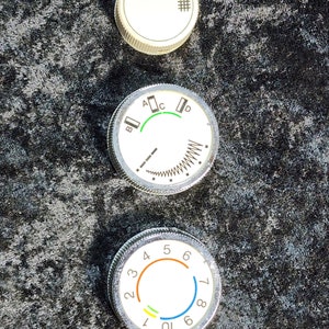 May include: Three round, silver-colored knobs with white faces and black markings. The top knob has a small grid pattern. The middle knob has a green arc and the letters A, B, C, and D. The bottom knob has numbers 1 through 10 and a colored arc with orange, yellow, and blue sections.