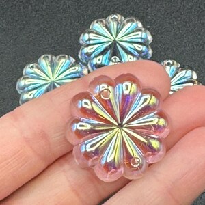 May include: Close-up of several iridescent flower-shaped beads. The beads have a pink hue with a shimmering, multi-colored effect. Each bead has a central hole and a petal design, perfect for jewelry making.