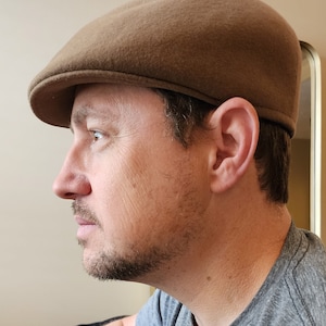 May include: A man wearing a brown felt flat cap. The cap has a slightly curved brim and a smooth surface.
